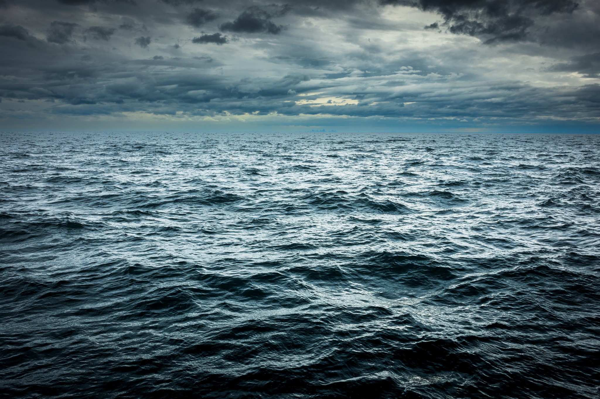 A vast expanse of choppy, dark blue ocean water under a heavy, dramatic overcast sky, with the faint silhouette of a distant city skyline barely visible on the horizon line.