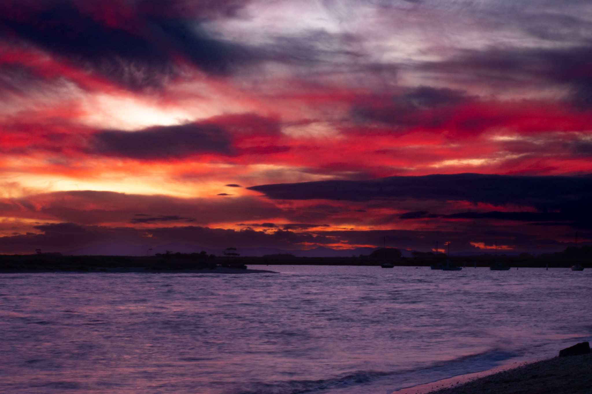 A dramatic sunset over a wide bay, with intense crimson, red, and purple clouds filling the sky and reflecting softly onto the rippling water below.